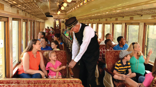 A vintage train interior with passengers of all ages enjoying the ride, while a conductor interacts with a young girl.