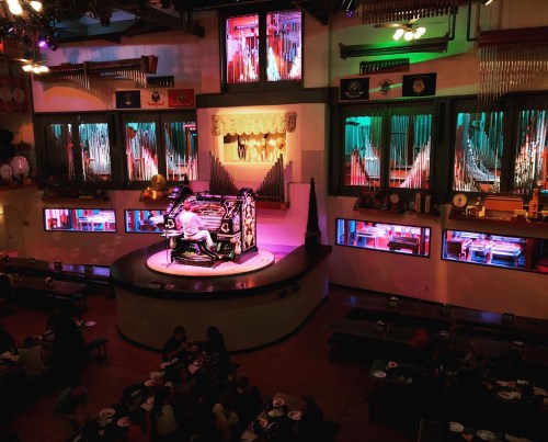A vibrant performance space with a large organ, colorful lights, and diners enjoying a show in a lively atmosphere.