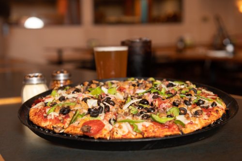 A large pizza topped with pepperoni, vegetables, and olives, served with a glass of beer and condiments on a table.
