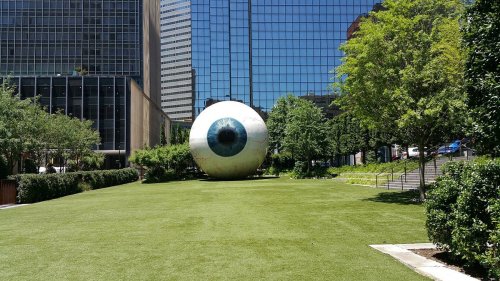 A large, surreal eyeball sculpture sits on a grassy area surrounded by trees and modern buildings.