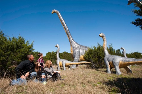 A group of people sits on the grass, gazing up at large dinosaur sculptures under a clear blue sky.