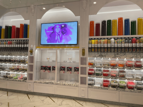 A colorful candy store display with jars of sweets, a large screen, and vibrant candy containers on the wall.