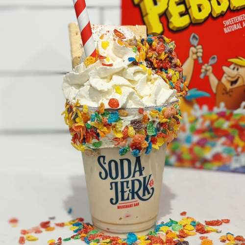 A colorful milkshake topped with whipped cream, cereal, and a cookie, served in a branded cup.