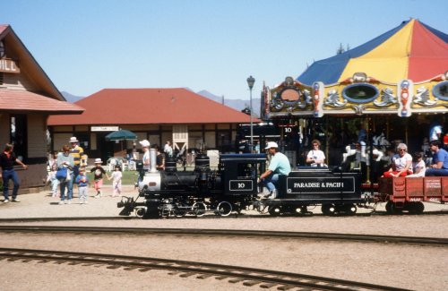 A miniature train rides past a carousel and people at a park on a sunny day.