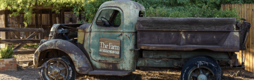 Rusty vintage truck with a wooden sign reading "The Farm" parked in a rustic outdoor setting.