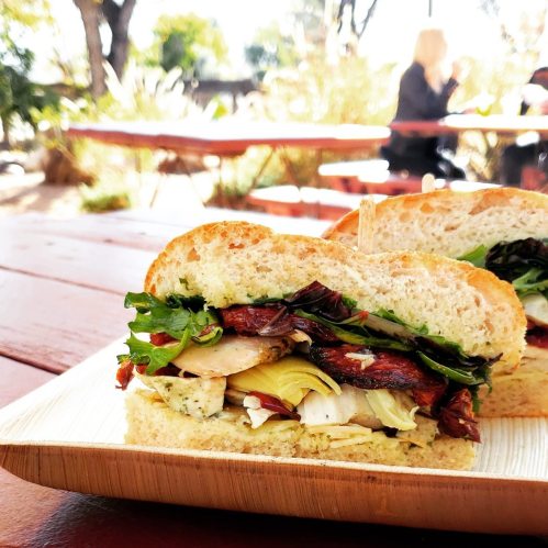 A close-up of a sandwich with layers of greens, artichokes, and sun-dried tomatoes on a wooden plate, outdoors.