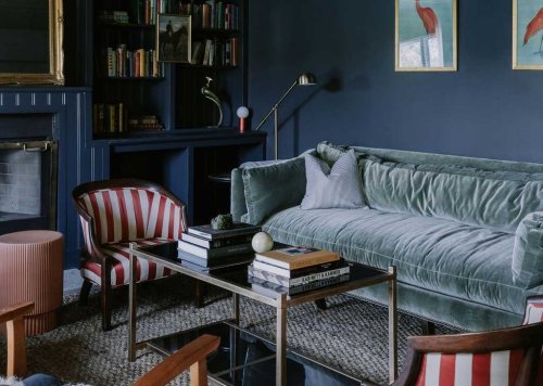 A cozy living room featuring a green velvet sofa, striped chairs, a glass coffee table, and bookshelves against a dark wall.