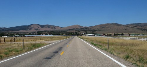 A straight, empty road stretches into the distance, flanked by fields and mountains under a clear blue sky.