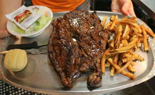 A large grilled steak on a silver platter, accompanied by fries, a roll, and a small salad.
