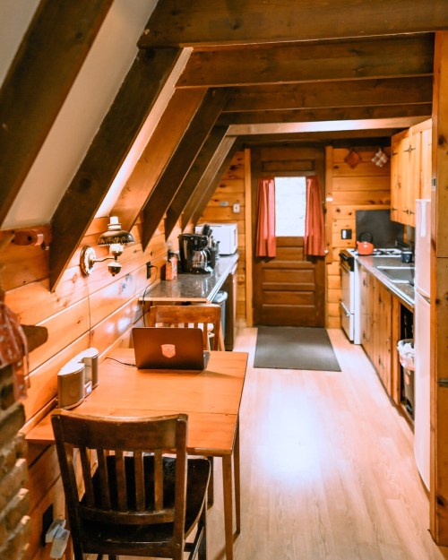 Cozy kitchen with wooden walls, a table with a laptop, and appliances, leading to a door with red curtains.