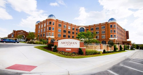 A modern brick building with domed roofs, featuring a sign that reads "Artesian," surrounded by landscaped greenery.
