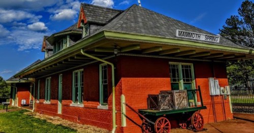 Historic brick train station with green accents, featuring a wooden cart outside, under a blue sky with clouds.