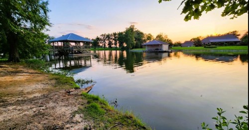 A serene lakeside view at sunset, featuring a dock and two buildings reflected in calm water.