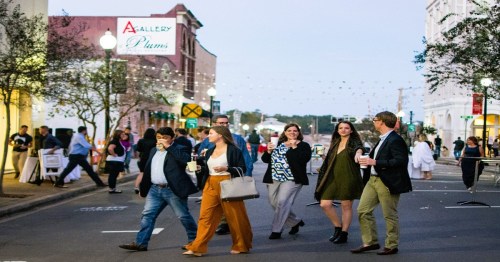 A group of people strolls down a lively street, enjoying the evening atmosphere and holding drinks.