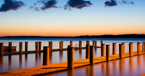 A serene sunset over a calm lake, with wooden docks and posts reflecting in the water.