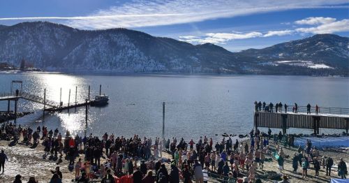 A crowded lakeside scene with people gathered on the shore and a dock, surrounded by snow-capped mountains and clear skies.