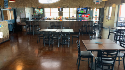 Interior of a casual restaurant with wooden tables, black chairs, a bar area, and a tropical decor theme.