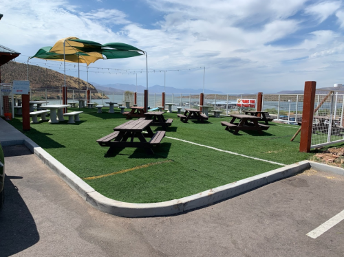 Outdoor seating area with picnic tables on artificial grass, shaded by a large canopy, overlooking a scenic view.