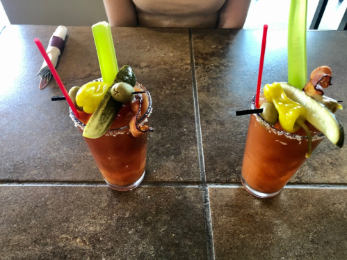 Two Bloody Mary cocktails garnished with celery, olives, pickles, and peppers on a table.
