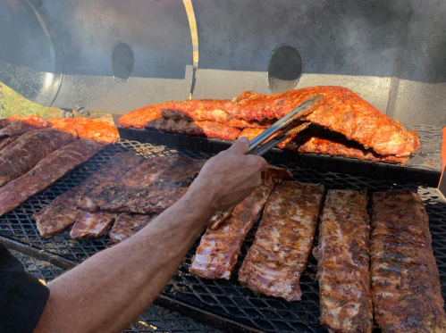 A hand using tongs to flip marinated ribs on a grill, with various cuts of meat cooking in the background.