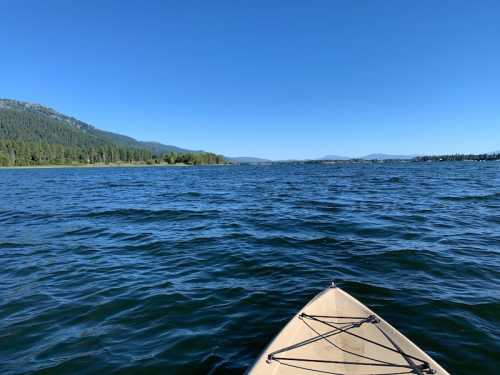 A kayak glides over calm blue water, with mountains and trees lining the shore under a clear blue sky.