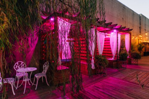 A cozy outdoor space with pink lighting, featuring wooden cabanas, white chairs, and lush greenery.
