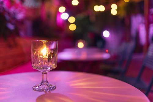 A lit candle in a glass sits on a table, surrounded by soft, colorful bokeh lights in a dimly lit setting.