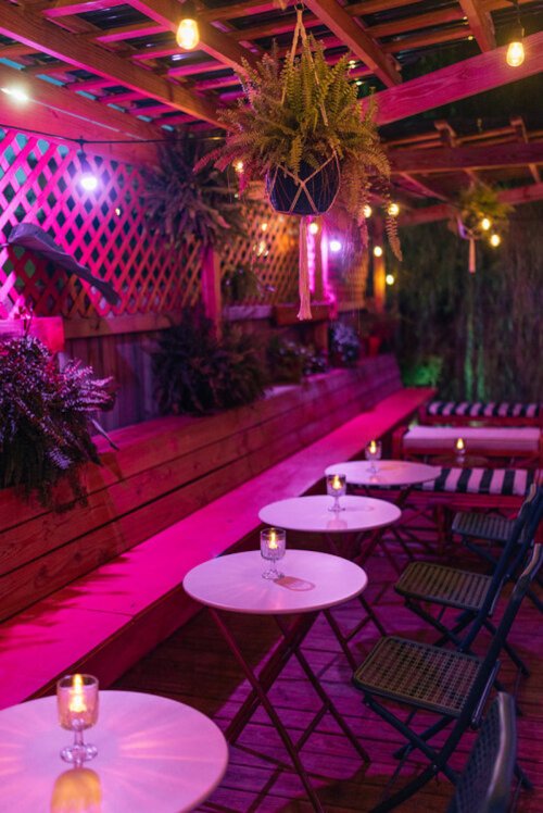 Cozy outdoor seating area with pink lighting, hanging plants, and candles on tables, creating a warm ambiance.