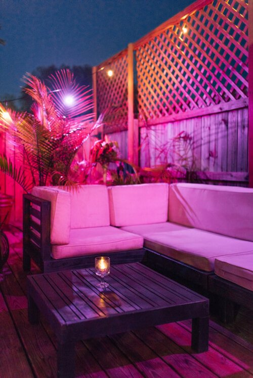 Cozy outdoor seating area with pink lighting, a wooden table, and plants, creating a warm evening ambiance.