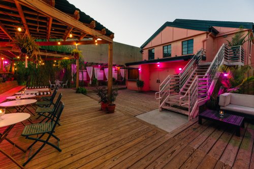 A cozy outdoor space with wooden decking, pink lighting, and seating areas surrounded by plants and a building in the background.