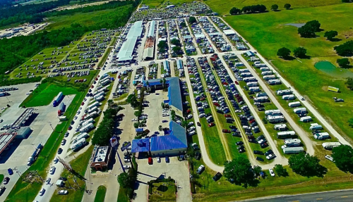 Aerial view of a large parking area filled with cars and RVs, surrounded by green fields and buildings.