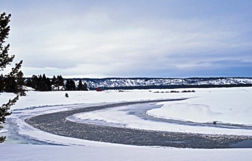 A winding road through a snowy landscape, with trees and distant hills under a cloudy sky.