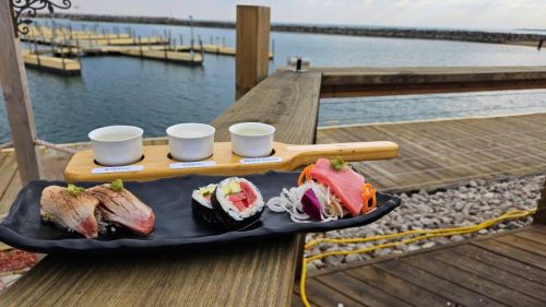A plate of sushi with three small cups of sauce, set against a scenic waterfront view.