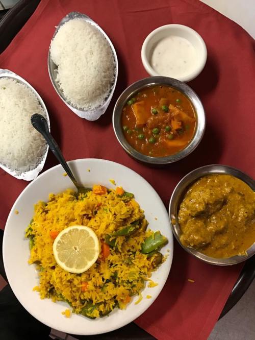 A colorful plate of biryani with lemon, accompanied by rice, curry, and yogurt on a red tablecloth.