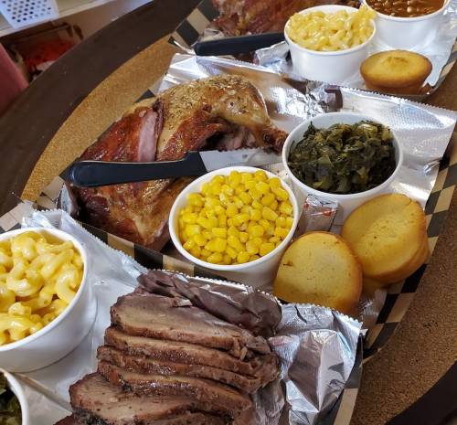 A platter of barbecue featuring sliced brisket, chicken, mac and cheese, corn, collard greens, and cornbread muffins.