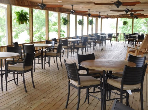 A spacious, empty outdoor dining area with wooden floors, round tables, and ceiling fans, surrounded by greenery.