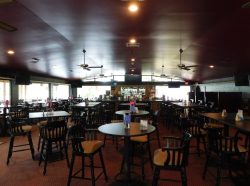 Interior of a spacious restaurant with dark wood tables, chairs, and a bar area, featuring ceiling fans and large windows.