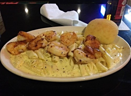 A plate of creamy pasta with grilled shrimp and a bread roll, served on a black table.