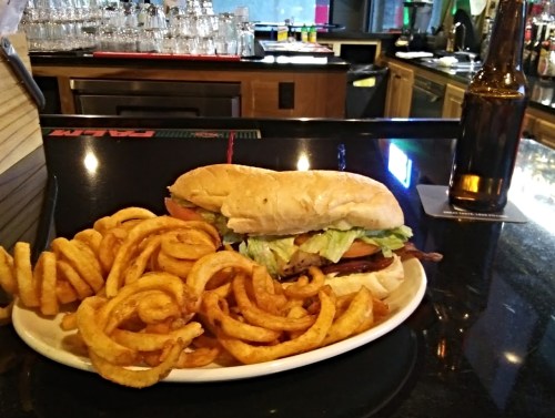 A sandwich with lettuce and tomato beside a plate of curly fries and a bottle of beer on a bar counter.