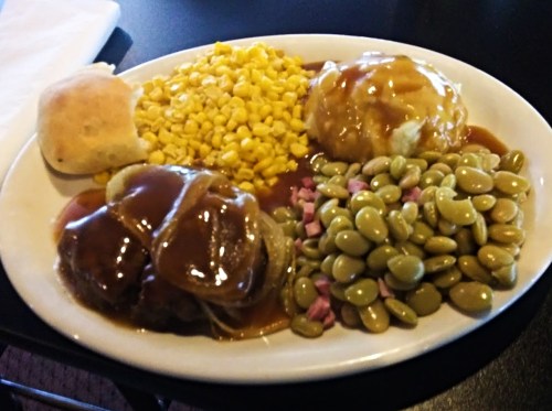 A plate of food featuring meatloaf with gravy, mashed potatoes, corn, and lima beans, alongside a biscuit.