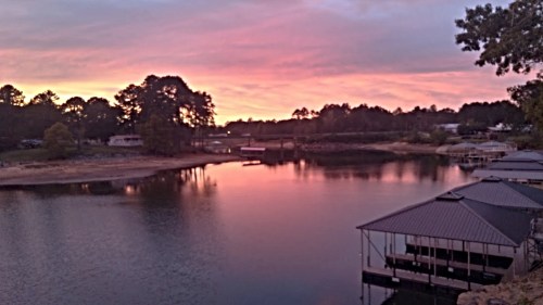 A serene lake at sunset, reflecting pink and orange hues, with docks and trees lining the shore.