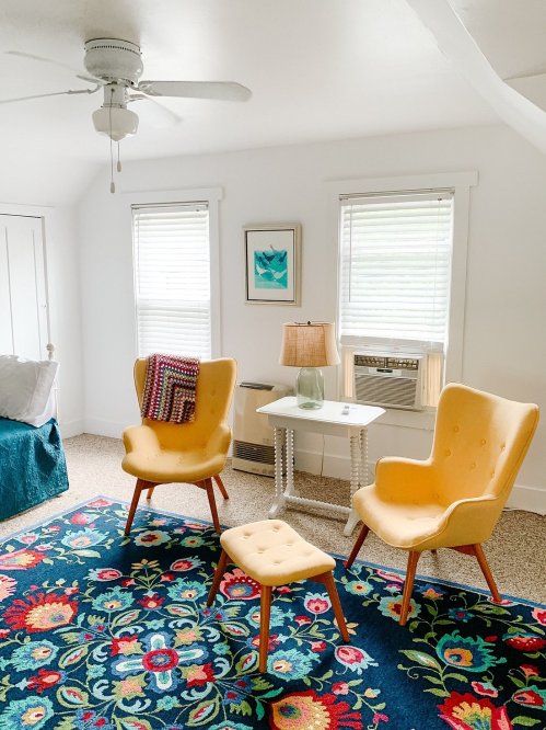Cozy room with yellow chairs, a small table, lamp, and colorful floral rug, featuring bright windows and a ceiling fan.
