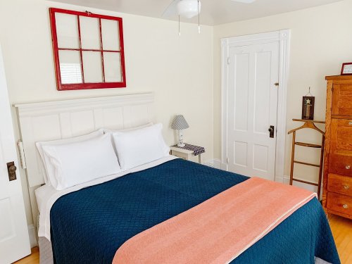 Cozy bedroom featuring a double bed with a blue and orange blanket, a wooden dresser, and a red-framed window.