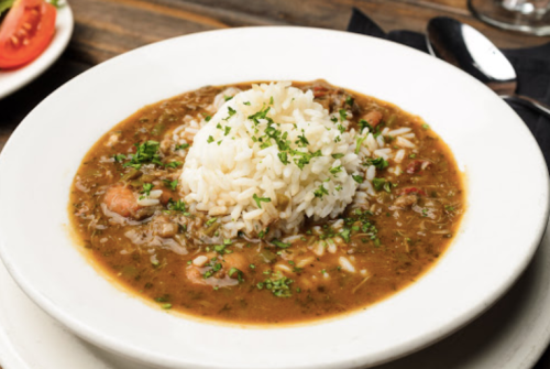A bowl of gumbo with rice and herbs, served on a wooden table.