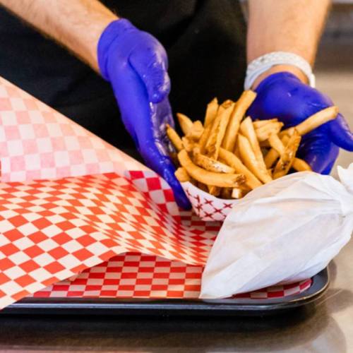 A person in blue gloves serves a bowl of golden fries on a checkered tray with red and white paper.