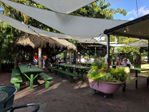 Outdoor dining area with green picnic tables, a thatched roof, and string lights, surrounded by lush greenery.