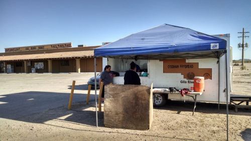 A blue tent set up in a parking lot, with two people inside and a sign for Gila River. Nearby buildings in the background.