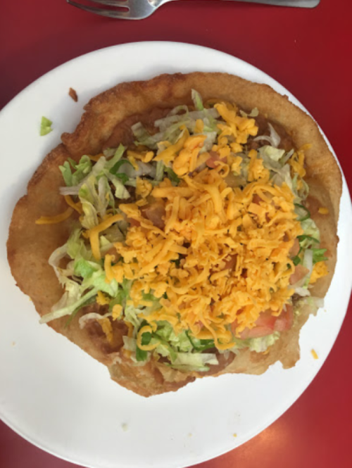 A crispy fried tortilla topped with lettuce, diced tomatoes, and shredded cheese on a white plate.