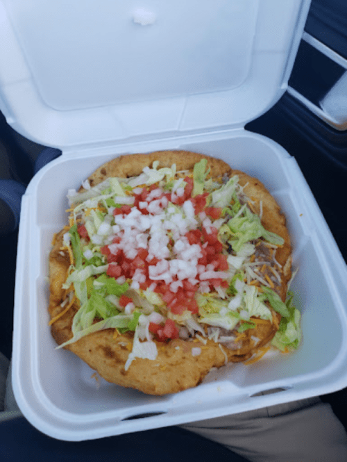 A takeout container holds a colorful taco salad topped with lettuce, tomatoes, onions, and cheese on a crispy shell.