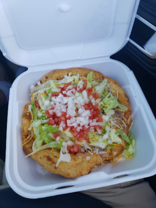 A takeout container holds a colorful taco salad topped with lettuce, tomatoes, onions, and cheese on a crispy shell.